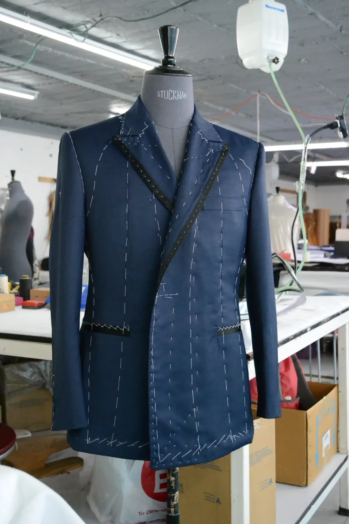 A half-made navy blazer with hand-basting stitches on a tailor's stand in Eddy's workshop