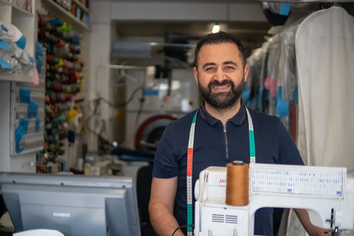 Eddy smiling in his dry cleaning and tailoring shop, tape measure around his neck