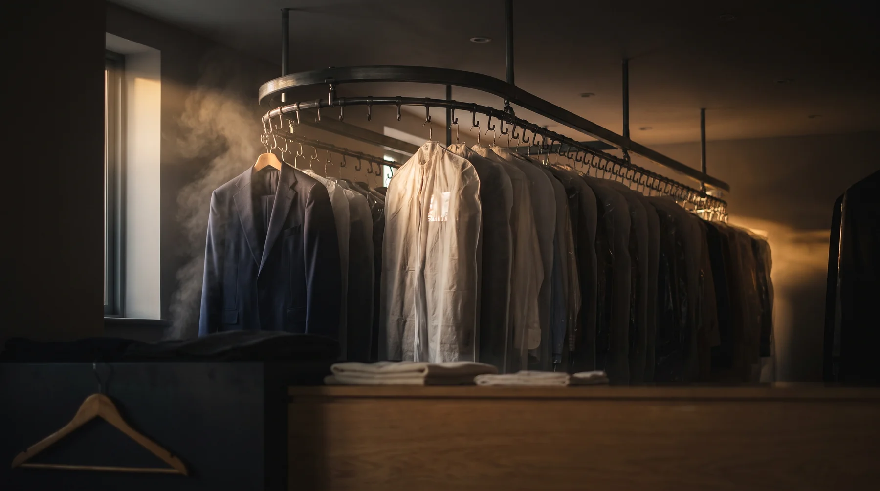 Moody interior of a dry-cleaning shop at dusk — a tailored navy suit and a rail of freshly cleaned garments in covers, steam catching warm low light